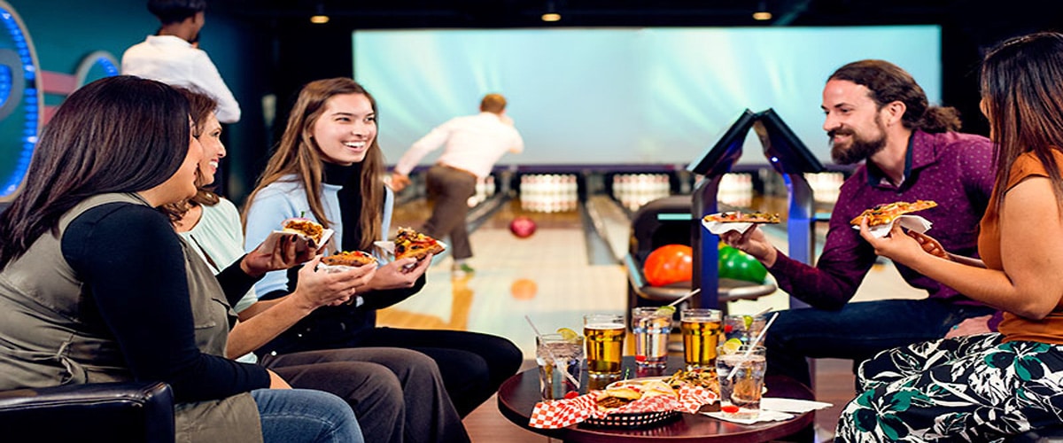 A group of friends enjoys pizza and drinks at a bowling lane table while someone bowls in the background.