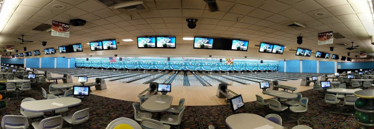 A wide panoramic view of a clean bowling alley with many lanes, overhead monitors, and table seating.