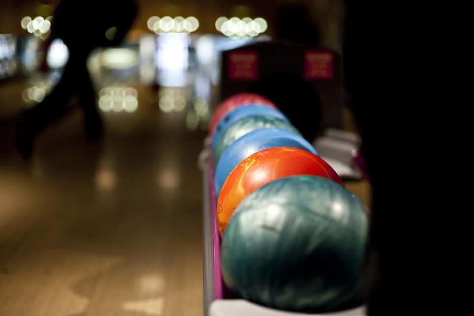 Close-up of colorful bowling balls on a rack with a blurred bowler and lights in the background.