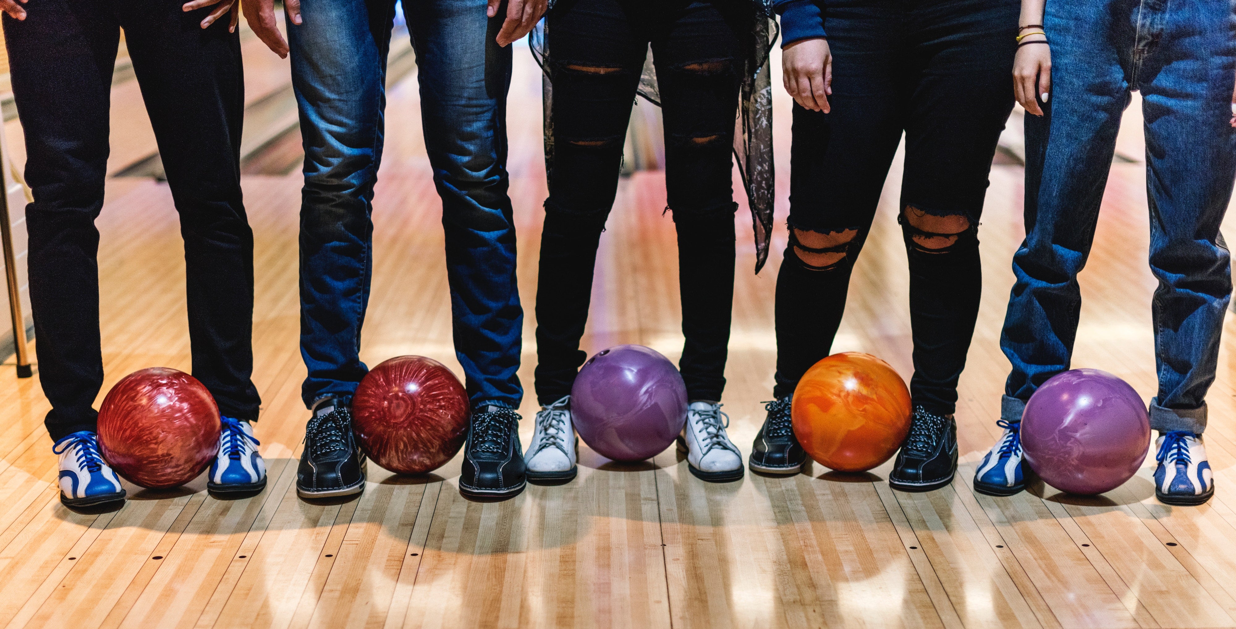 A waist-down view of five people standing on a wooden bowling lane with colorful bowling balls at their feet.