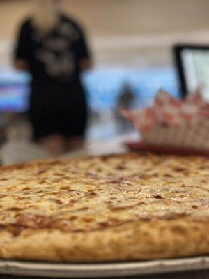 A close-up of a cheese pizza on a metal tray with a blurred bowling alley in the background.