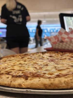 A close-up shot of a large cheese pizza on a metal tray with blurred bowling lanes in the background.
