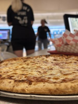 A fresh cheese pizza on a metal tray with a blurred bowling alley in the background.