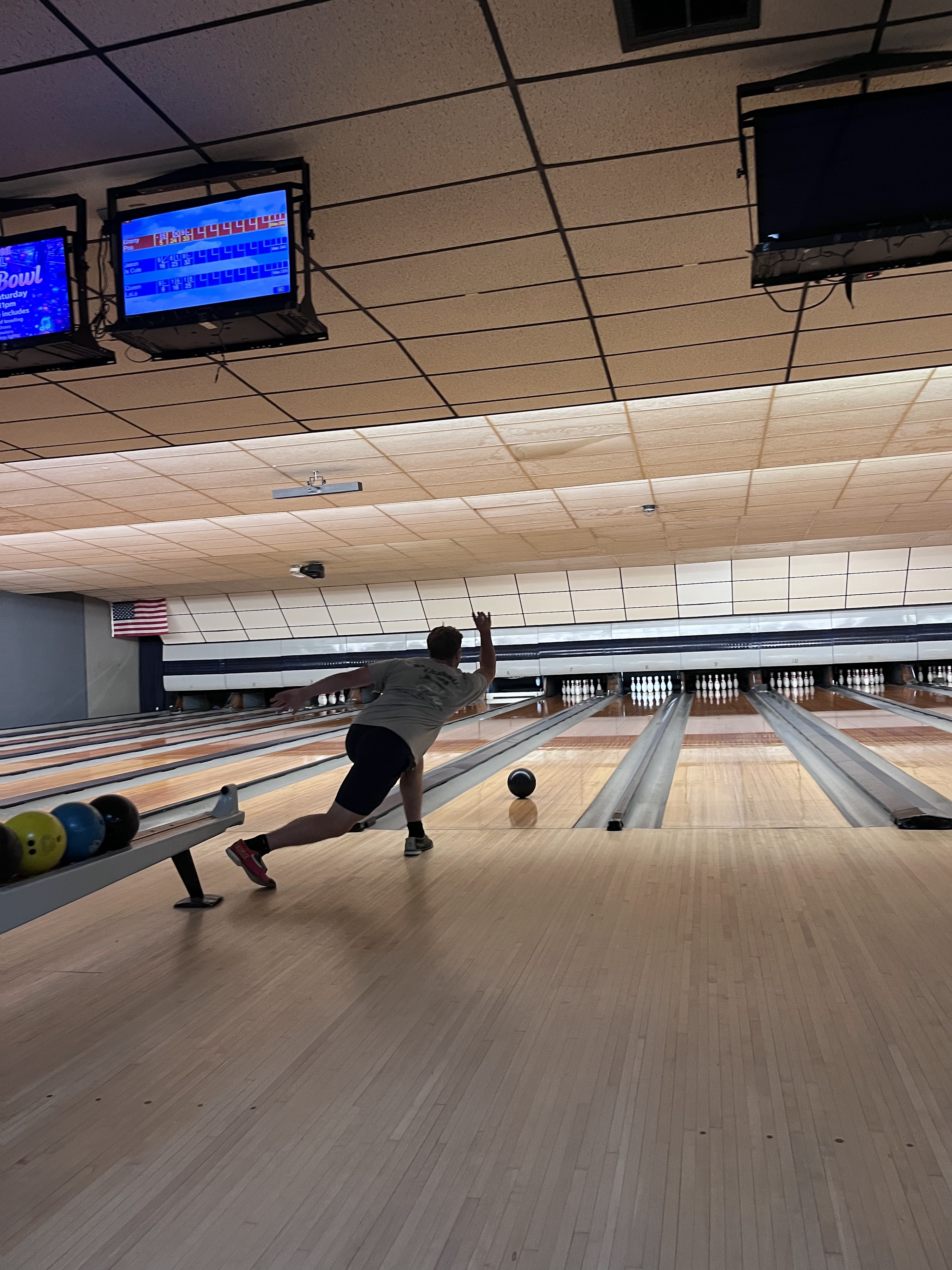 A man in a grey t-shirt releases a bowling ball on a wooden lane under overhead scoring monitors.