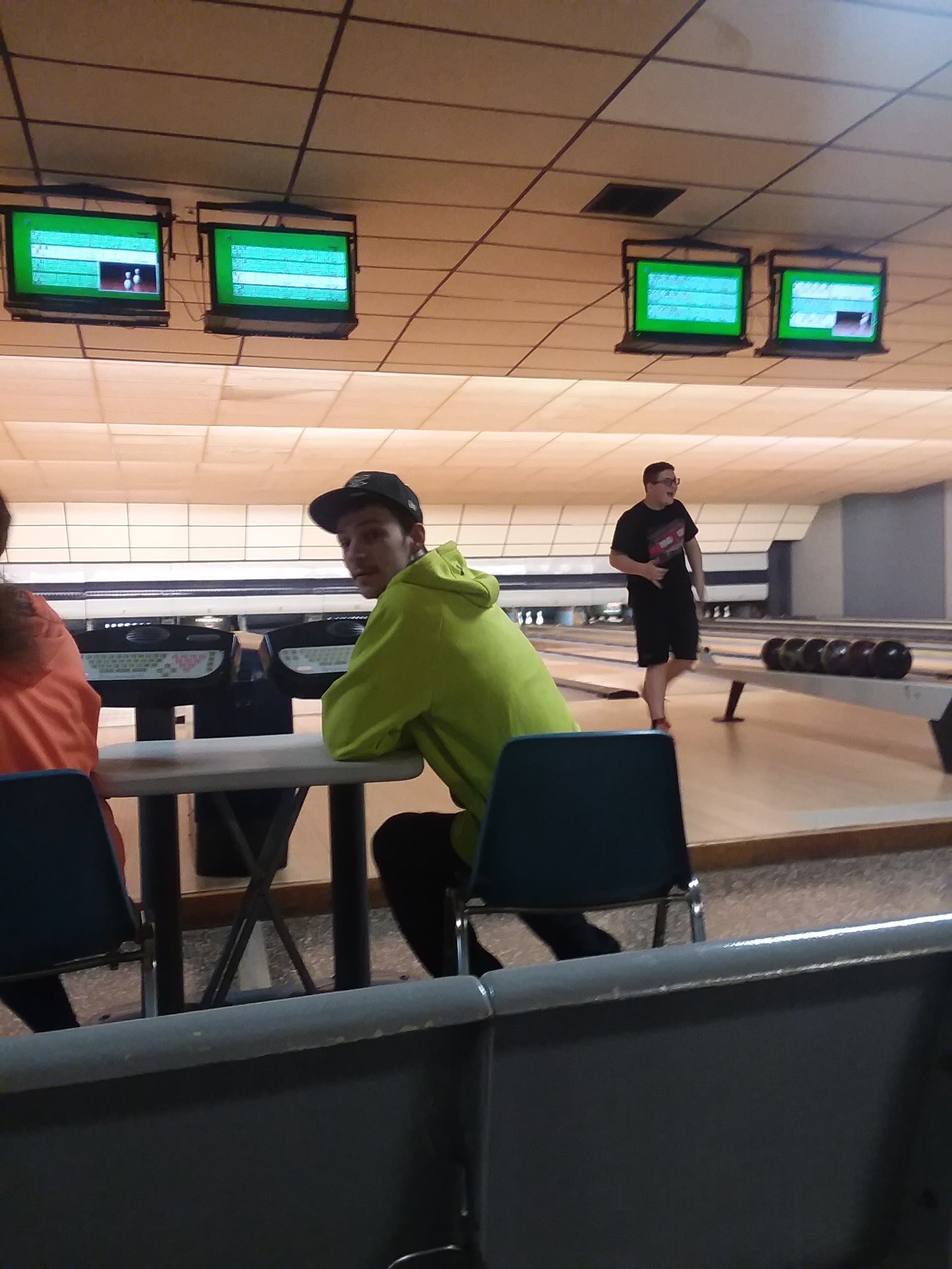 A person in a green hoodie sits at a bowling lane table with overhead scoring monitors in the background.