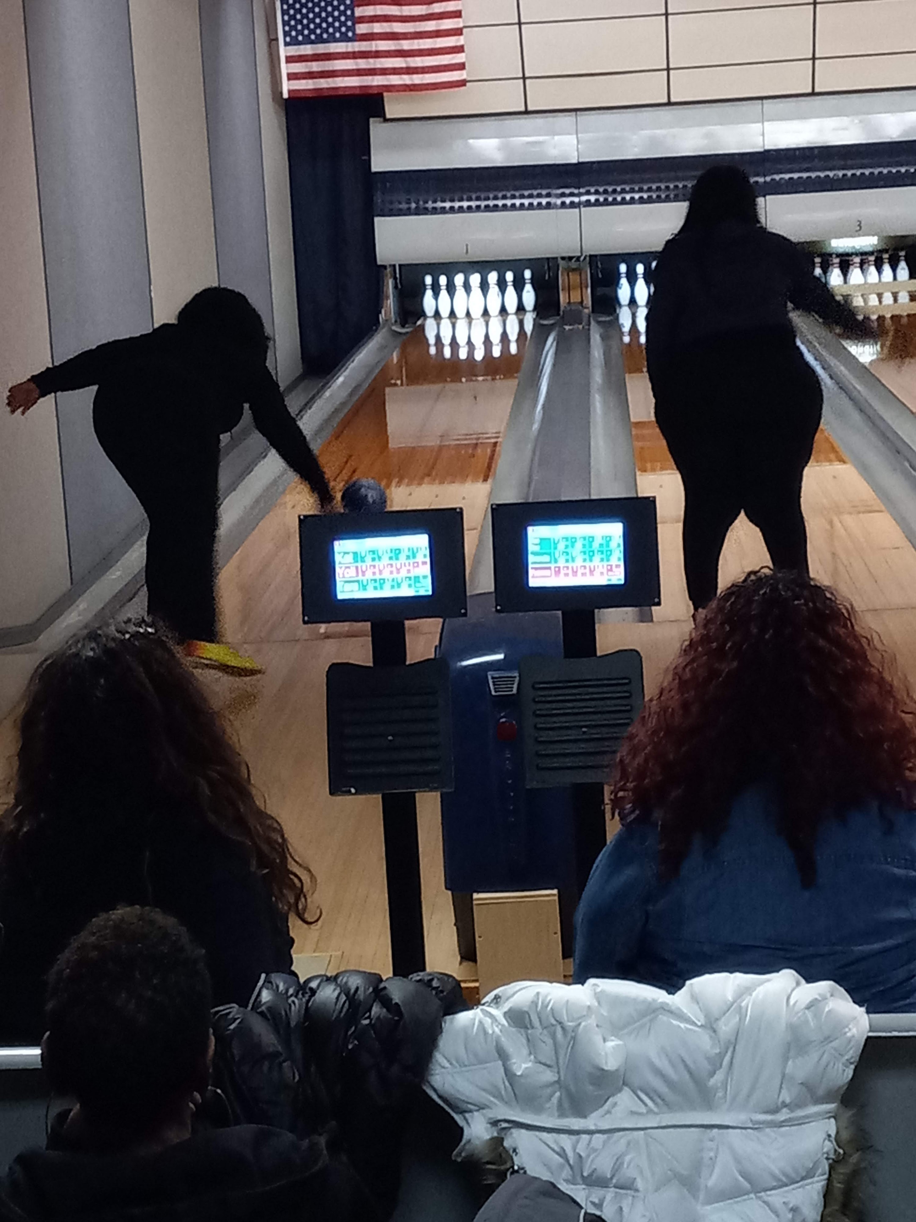 Two people bowling on hardwood lanes with digital scoring monitors while spectators watch from the foreground seats.