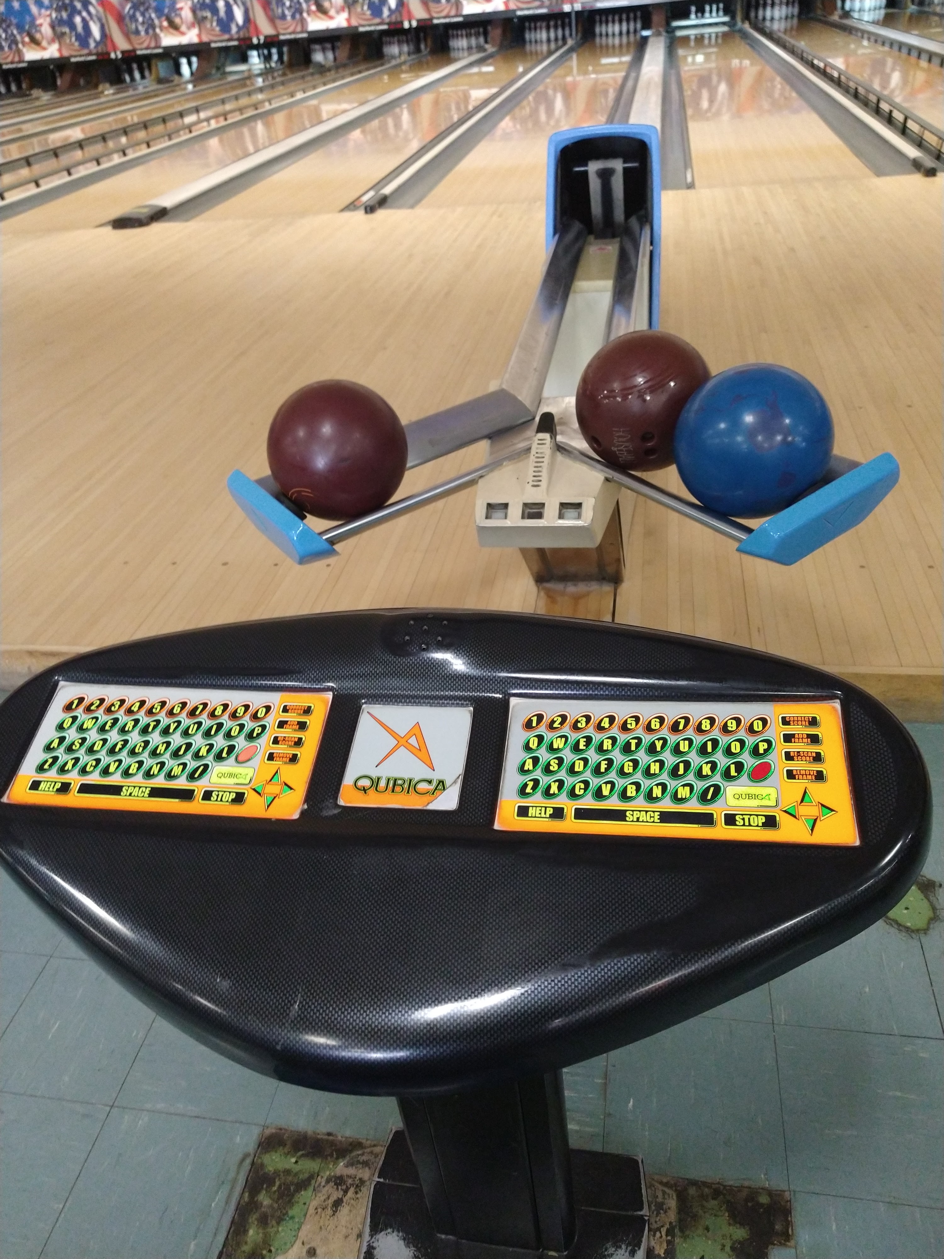A Qubica bowling scoring console and ball return with three bowling balls in front of several bowling lanes.