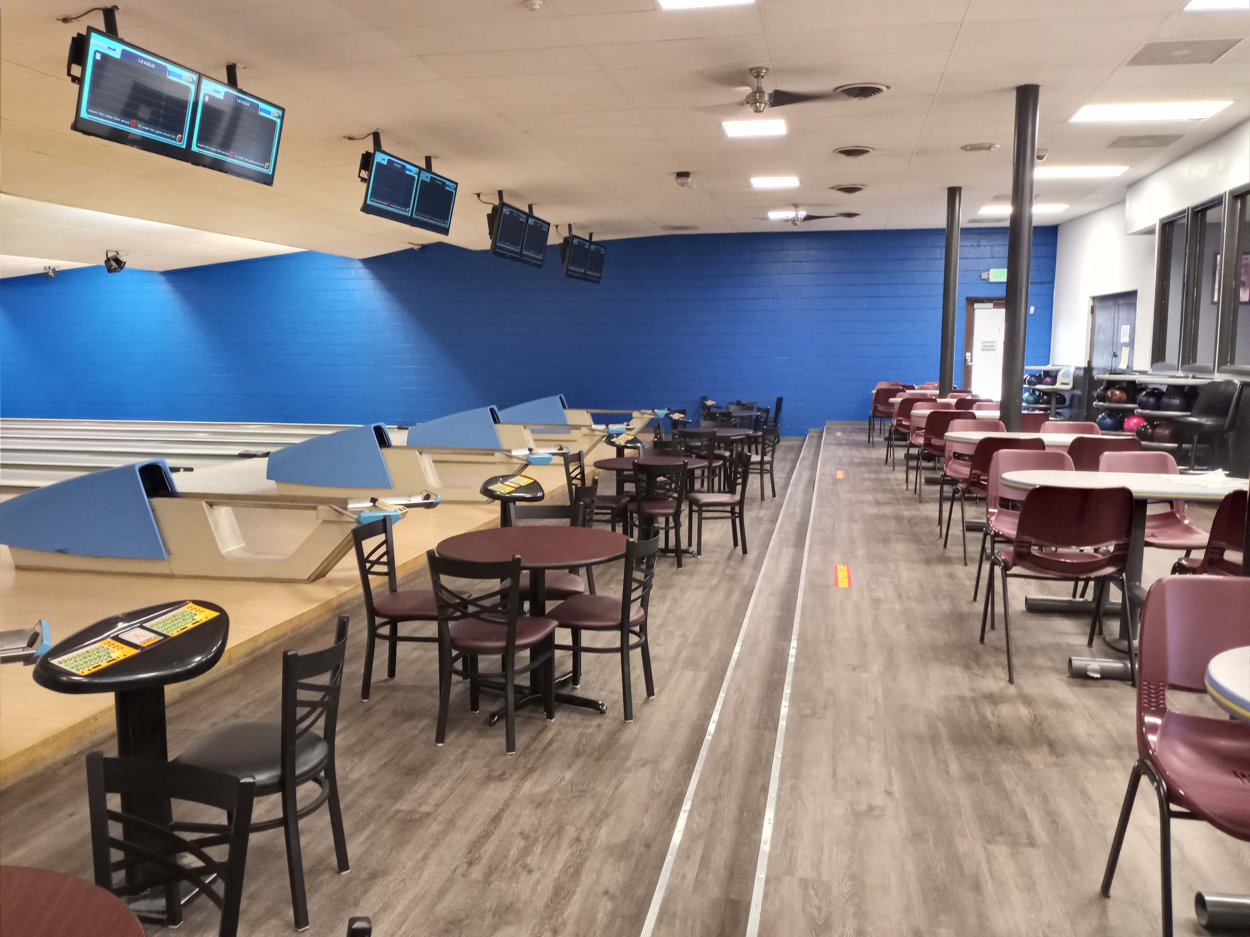 Wide view of bowling lanes with blue ball returns, overhead scoring monitors, and several rows of tables and chairs.