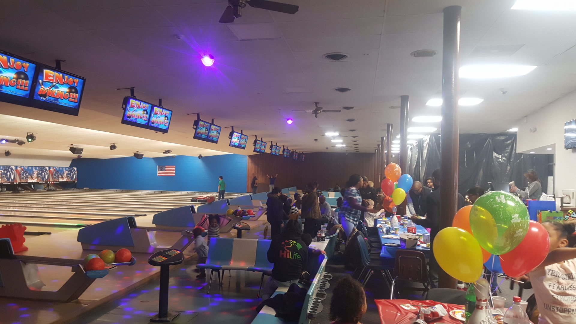 A colorful birthday party celebration with balloons and guests next to bowling lanes at an indoor center.