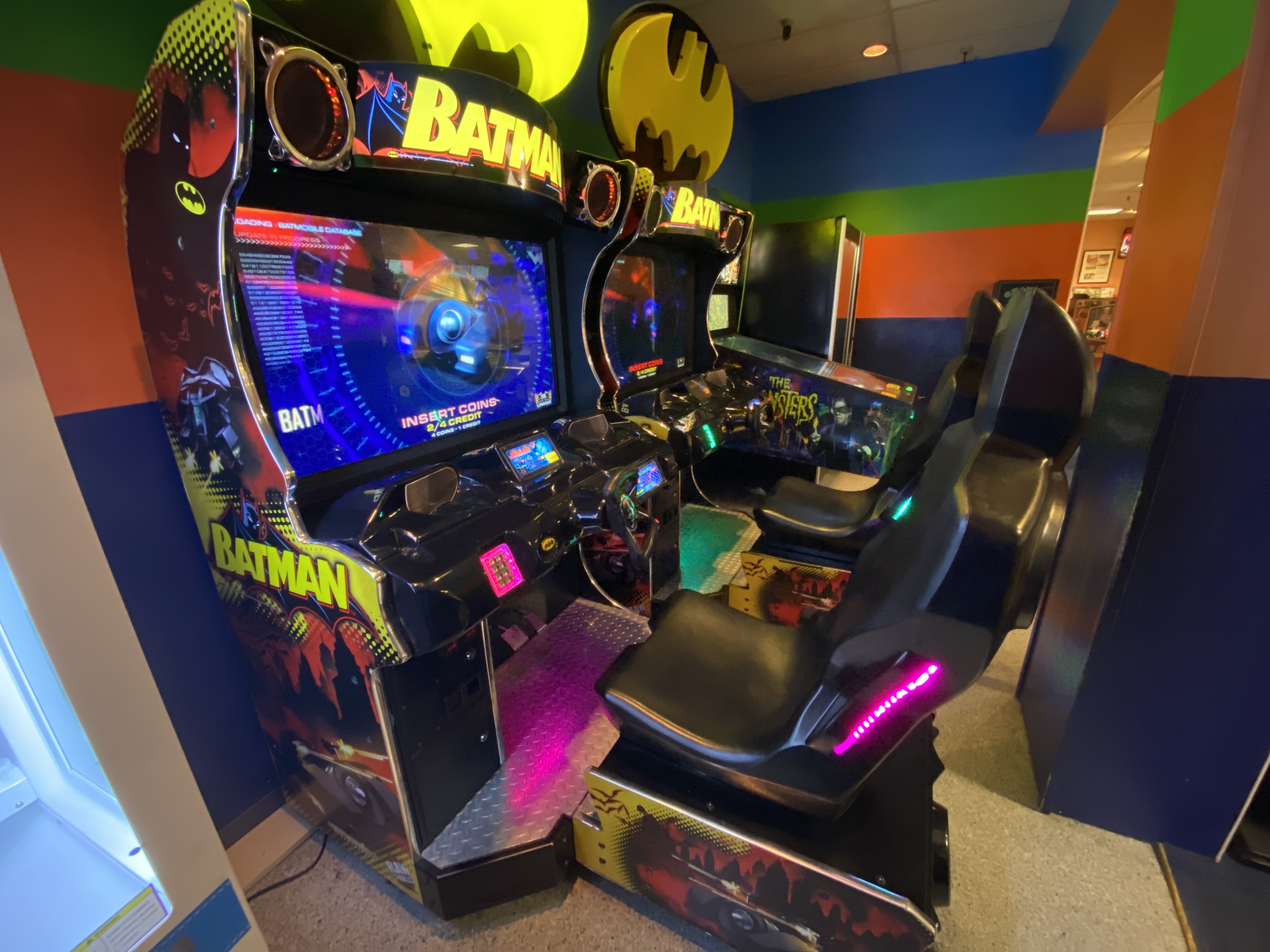 Two Batman-themed sit-down racing arcade games with yellow bat-signals on top in a room with colorful striped walls.