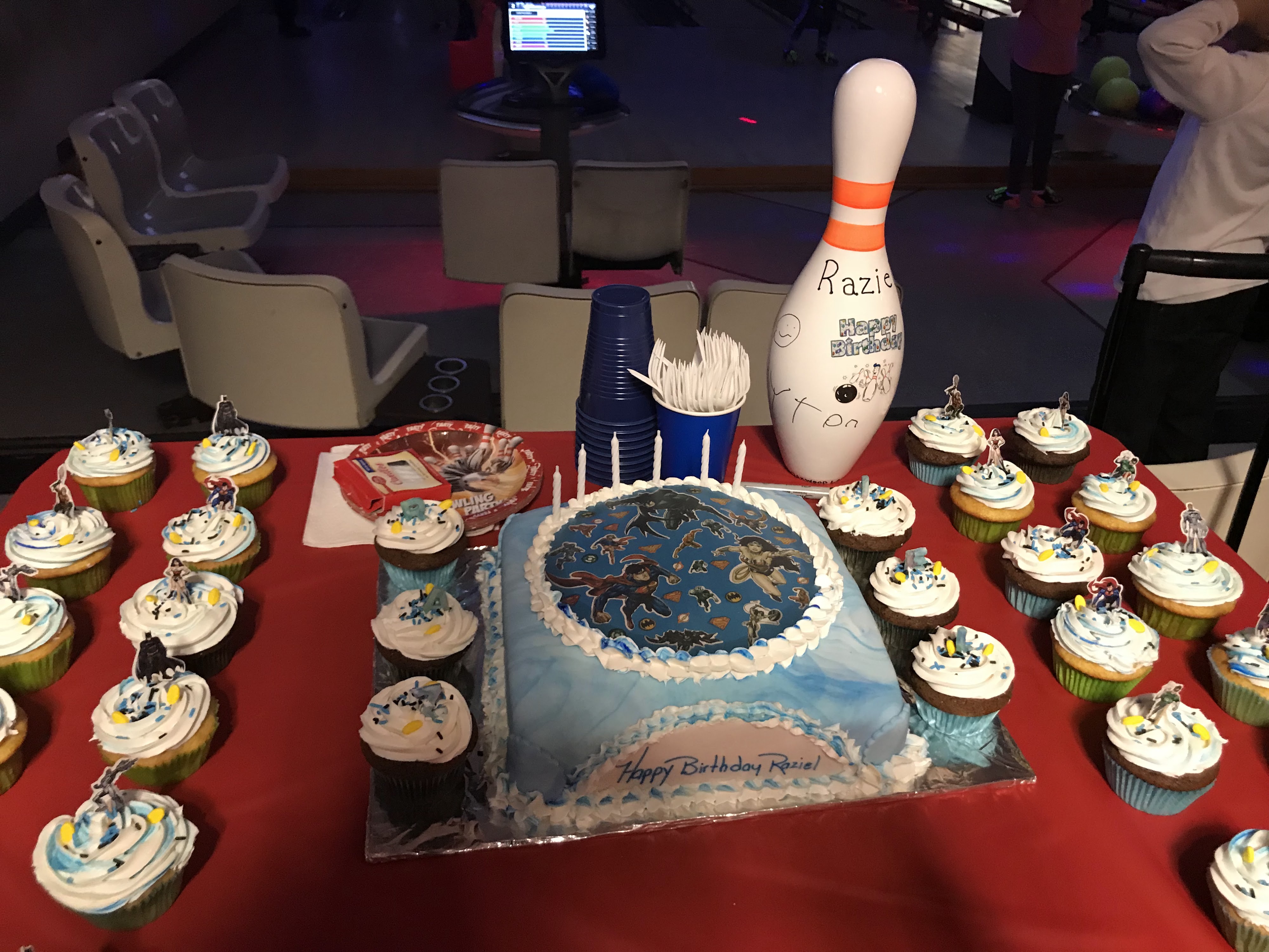 A superhero-themed birthday cake and cupcakes on a red table in front of bowling lanes with glow lighting.