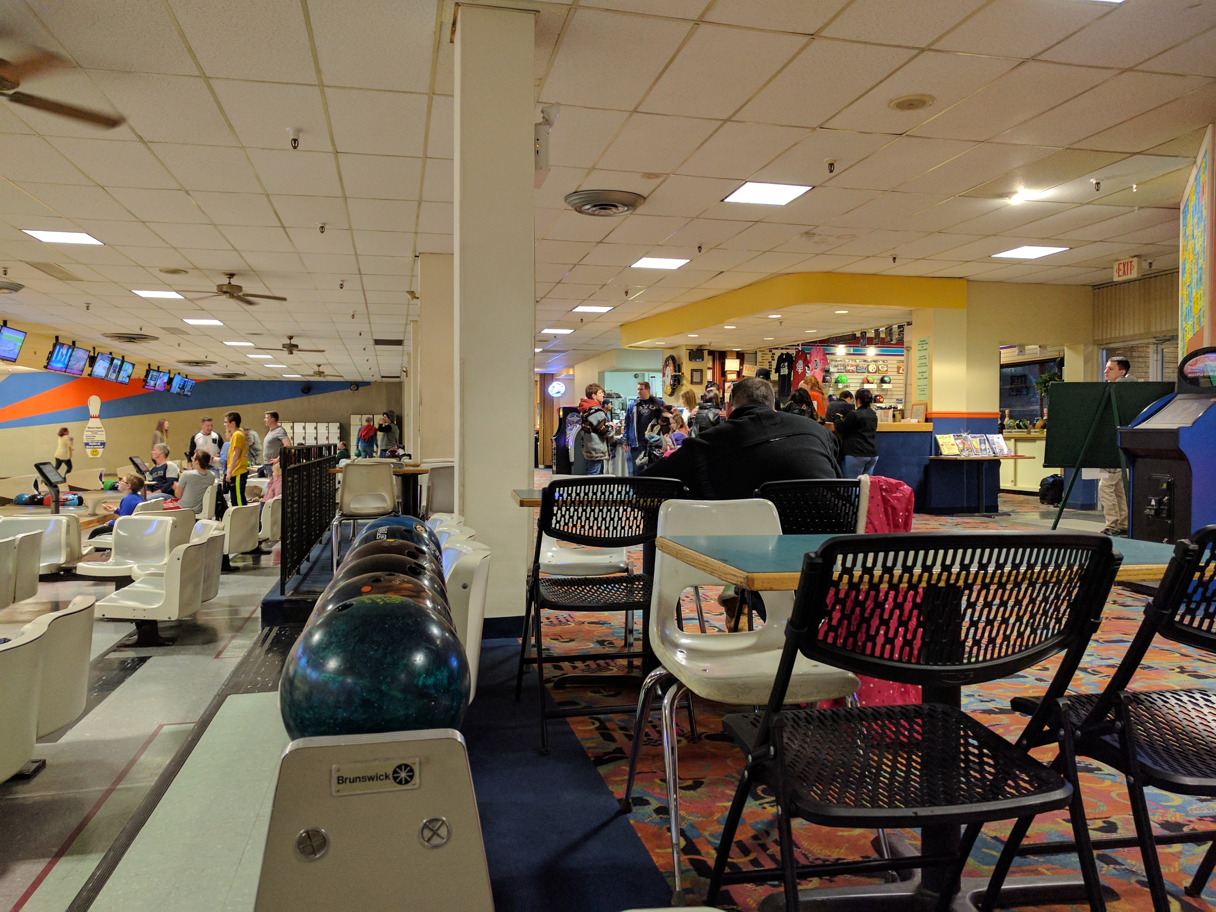 A busy interior view of a bowling center with ball racks, seating, and lanes visible in the background.