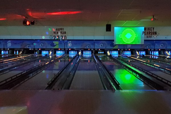Bowling lanes at night with colorful neon glow lighting and a projection screen.