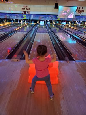 A young child uses an orange bowling ramp on a lane with colorful glow lighting and bumpers.