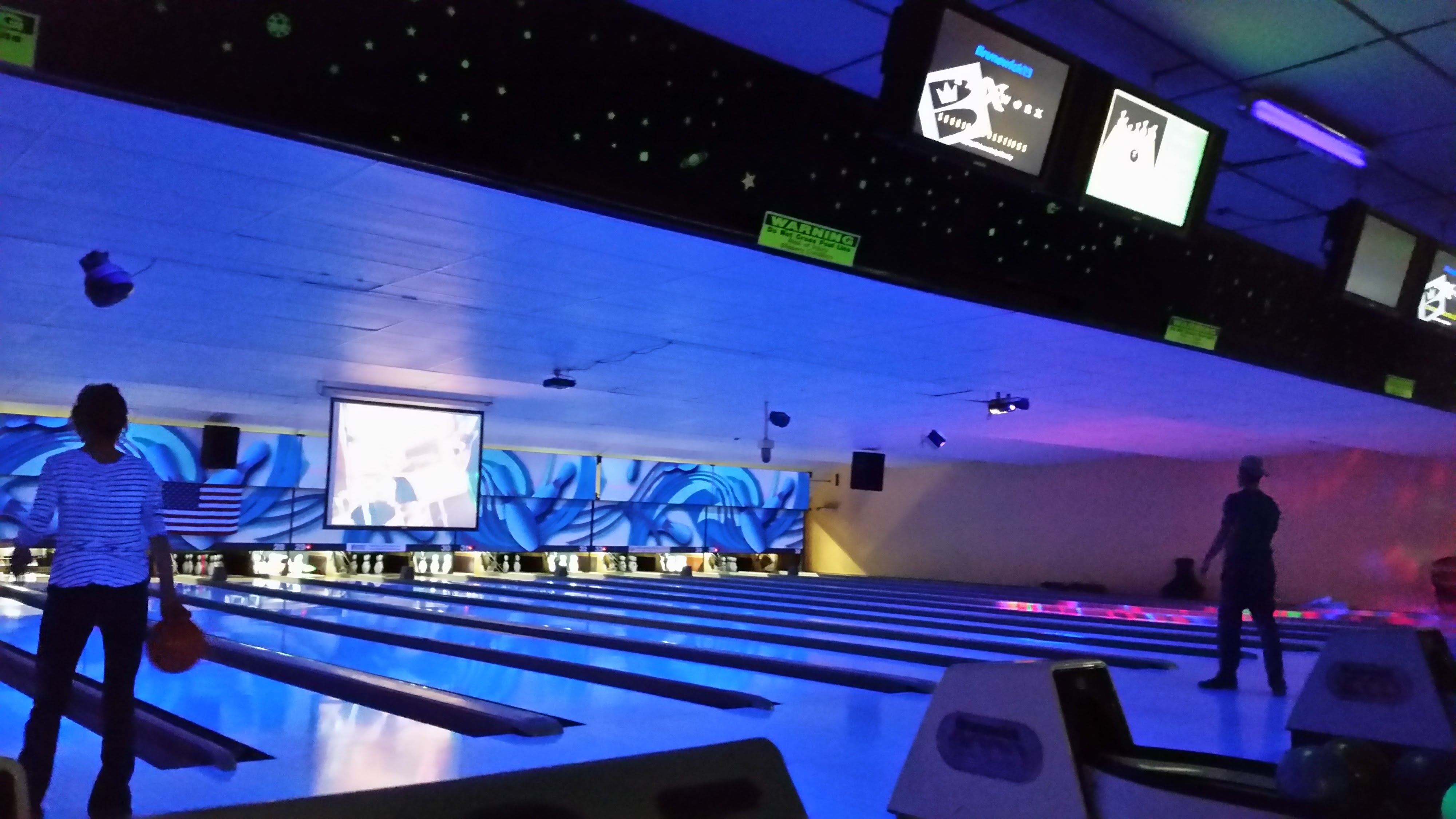 Bowling lanes under blue and purple cosmic glow lighting with players and overhead score monitors.