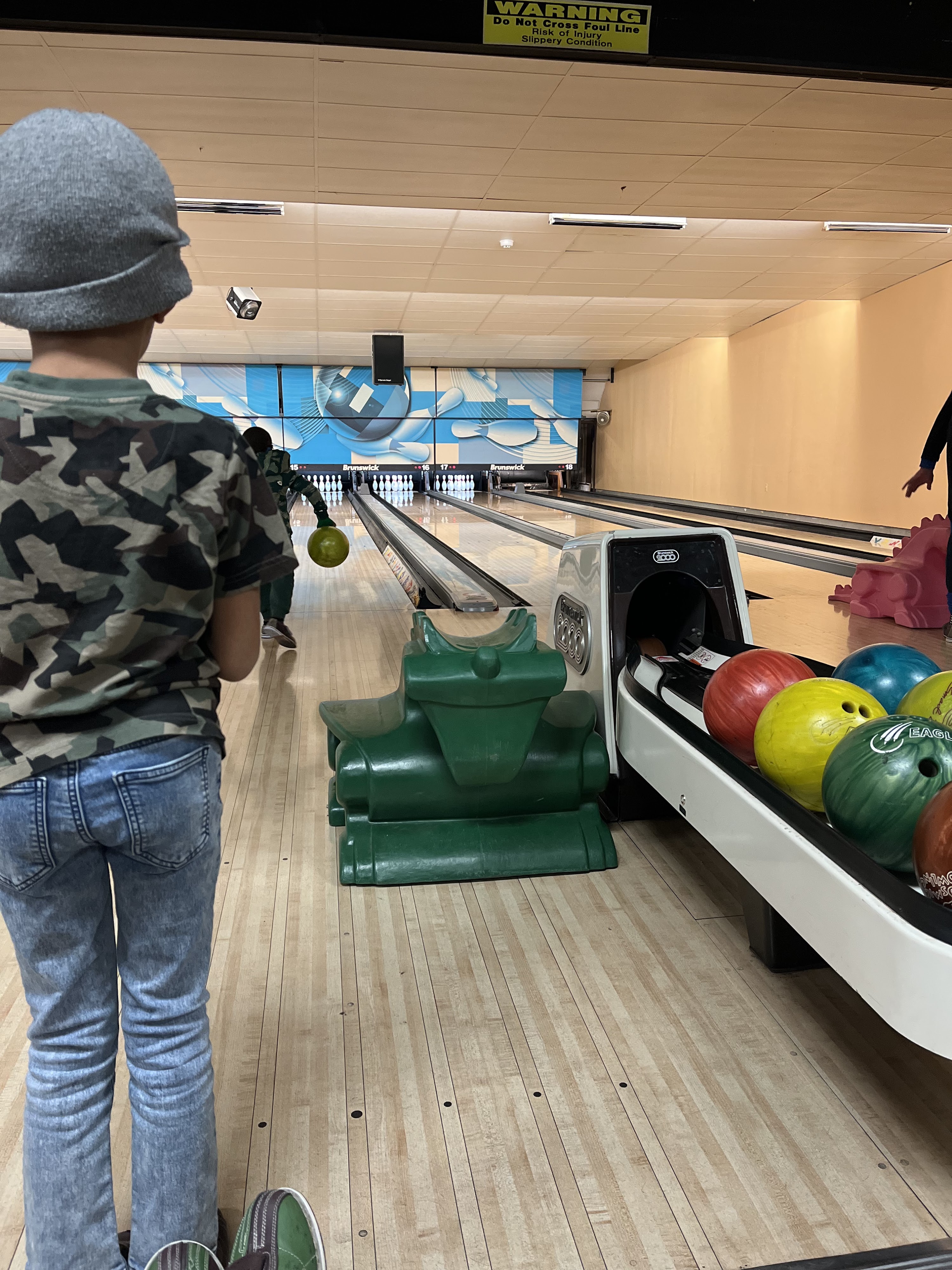 A young child watching another bowler on a lane equipped with a green dragon ramp and bumpers.