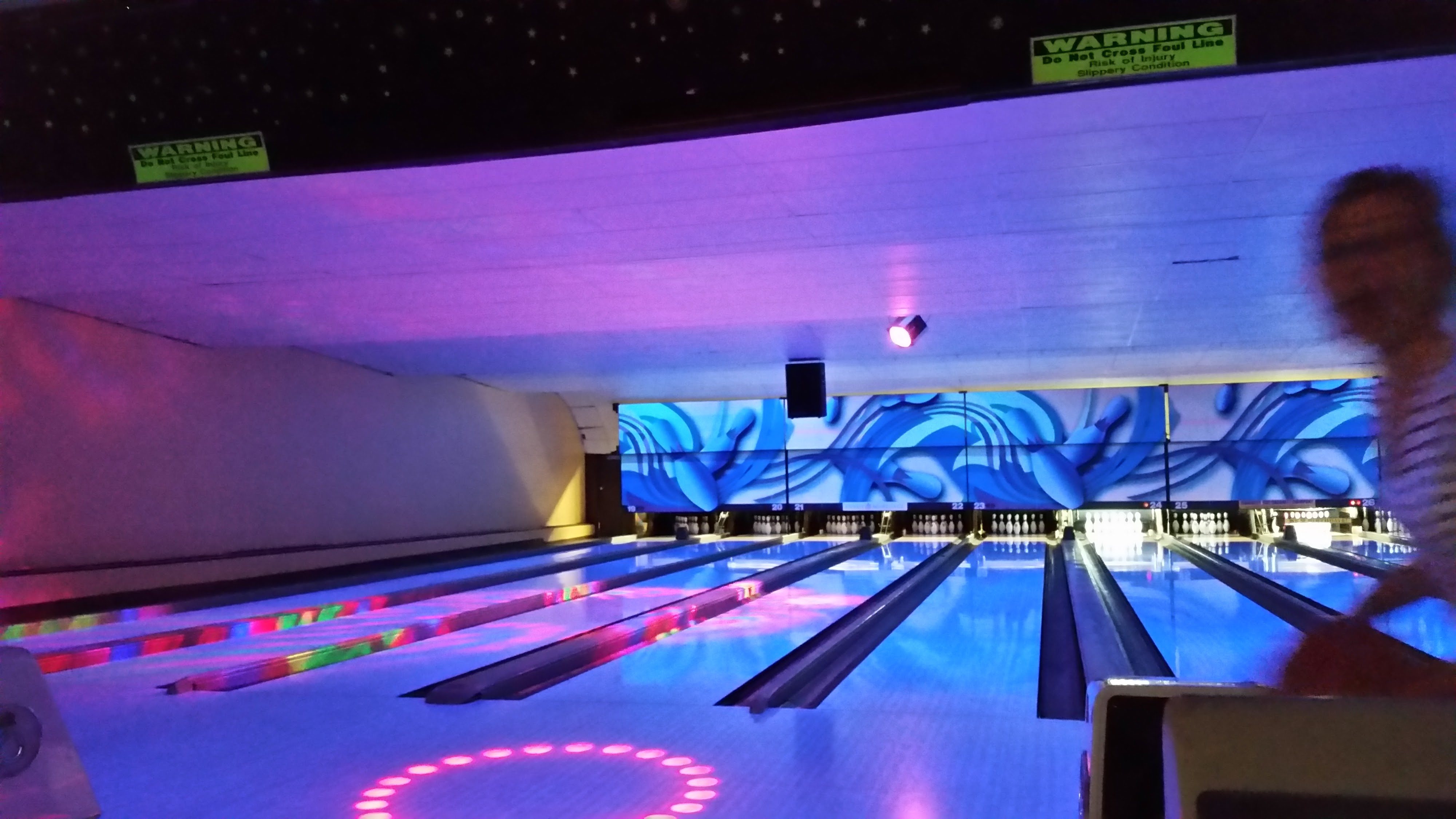 A bowling alley interior with lanes glowing under blue and purple cosmic lighting and neon floor patterns.