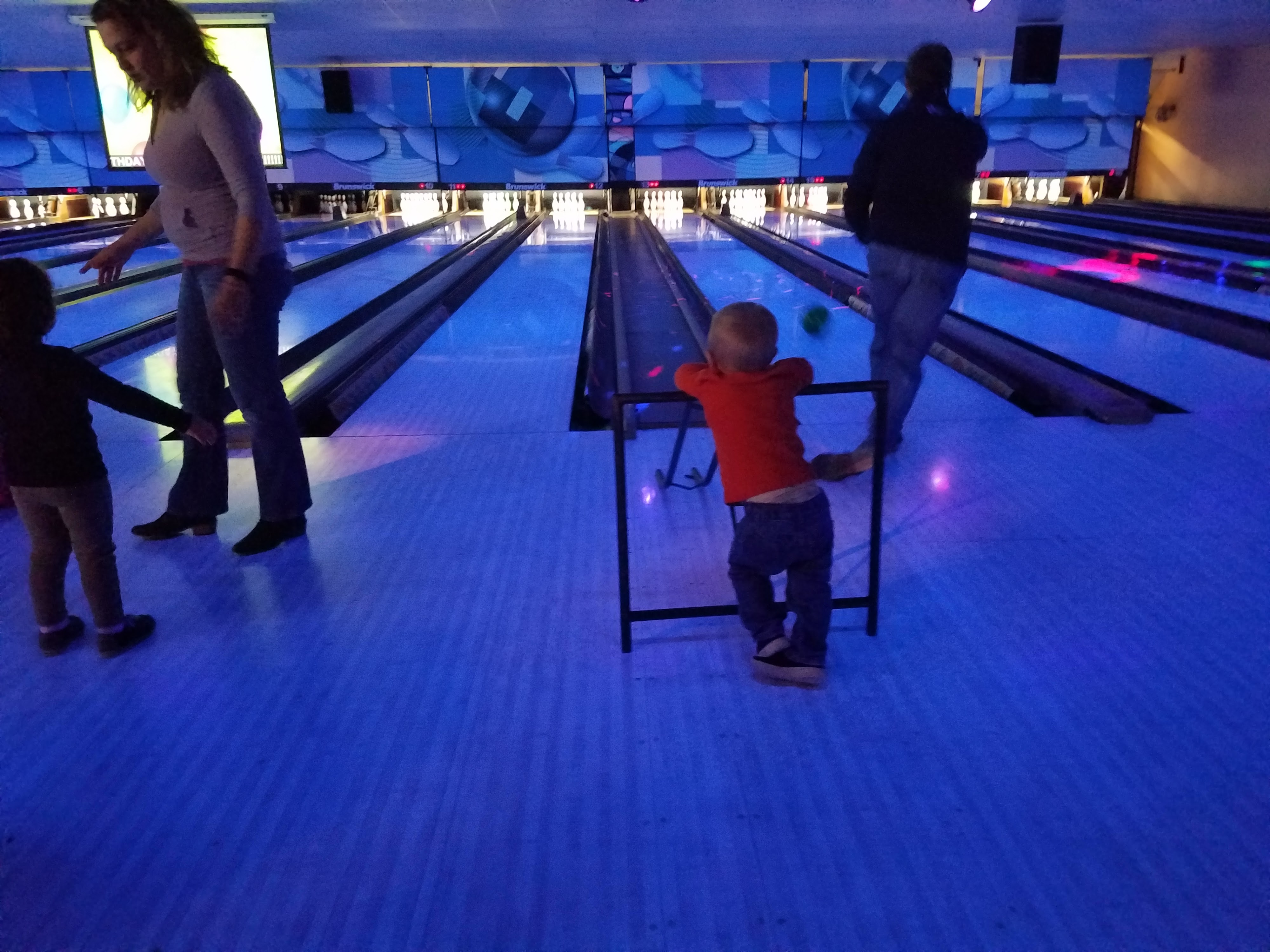 A family enjoys cosmic bowling with a young boy using a metal ball ramp on a blue-lit lane.