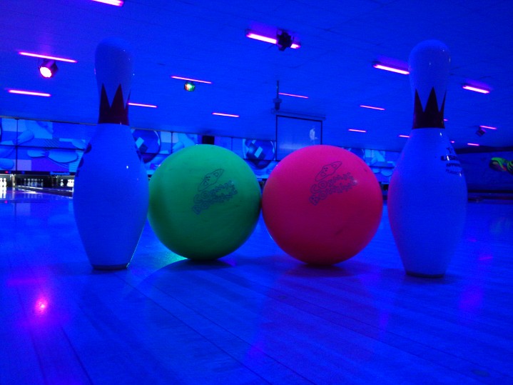 Two neon bowling balls and two pins on a lane under blue cosmic lighting.