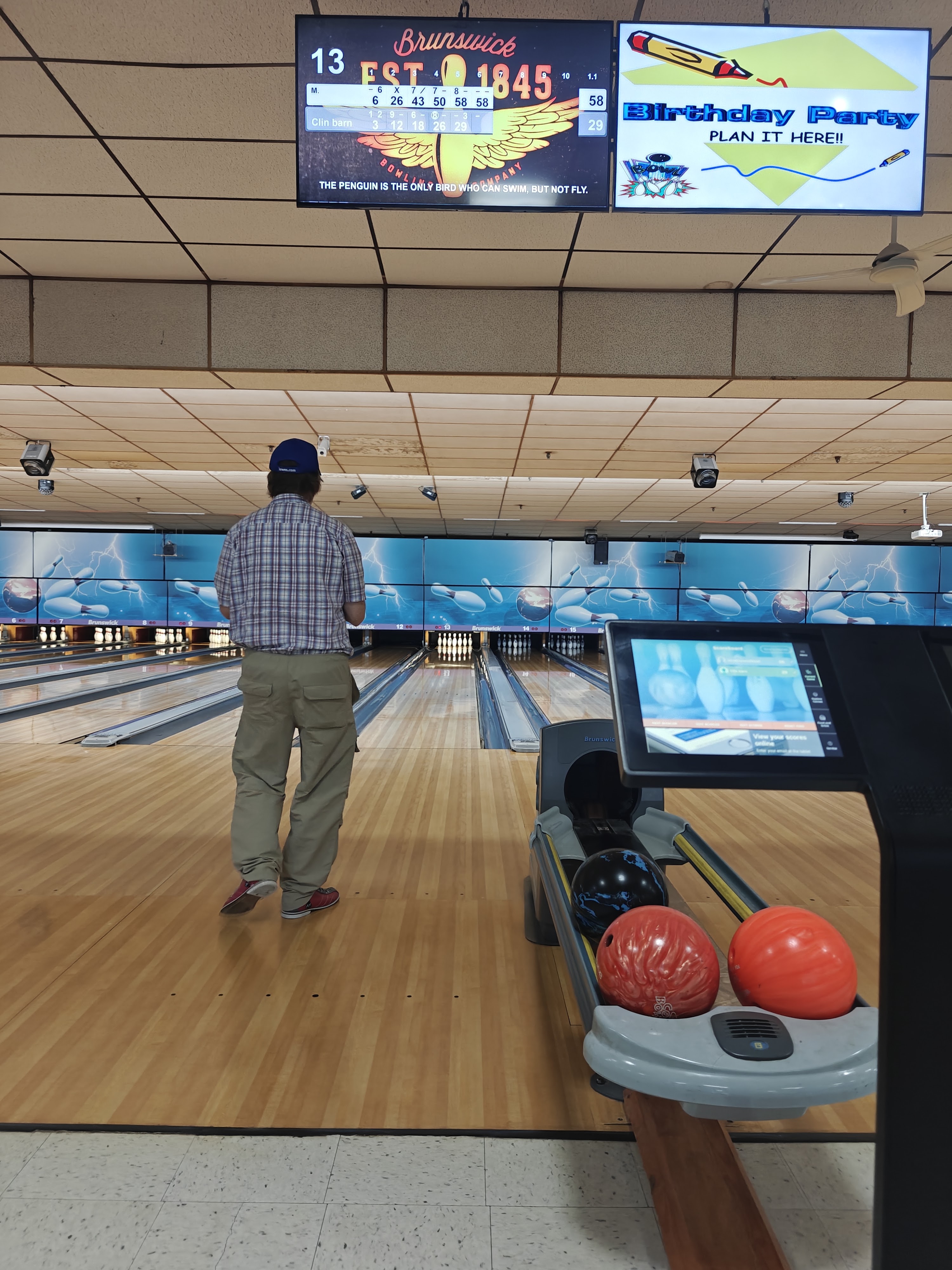 A bowler walks down a lane towards the pins, with bowling balls in the return and digital scoring screens overhead.
