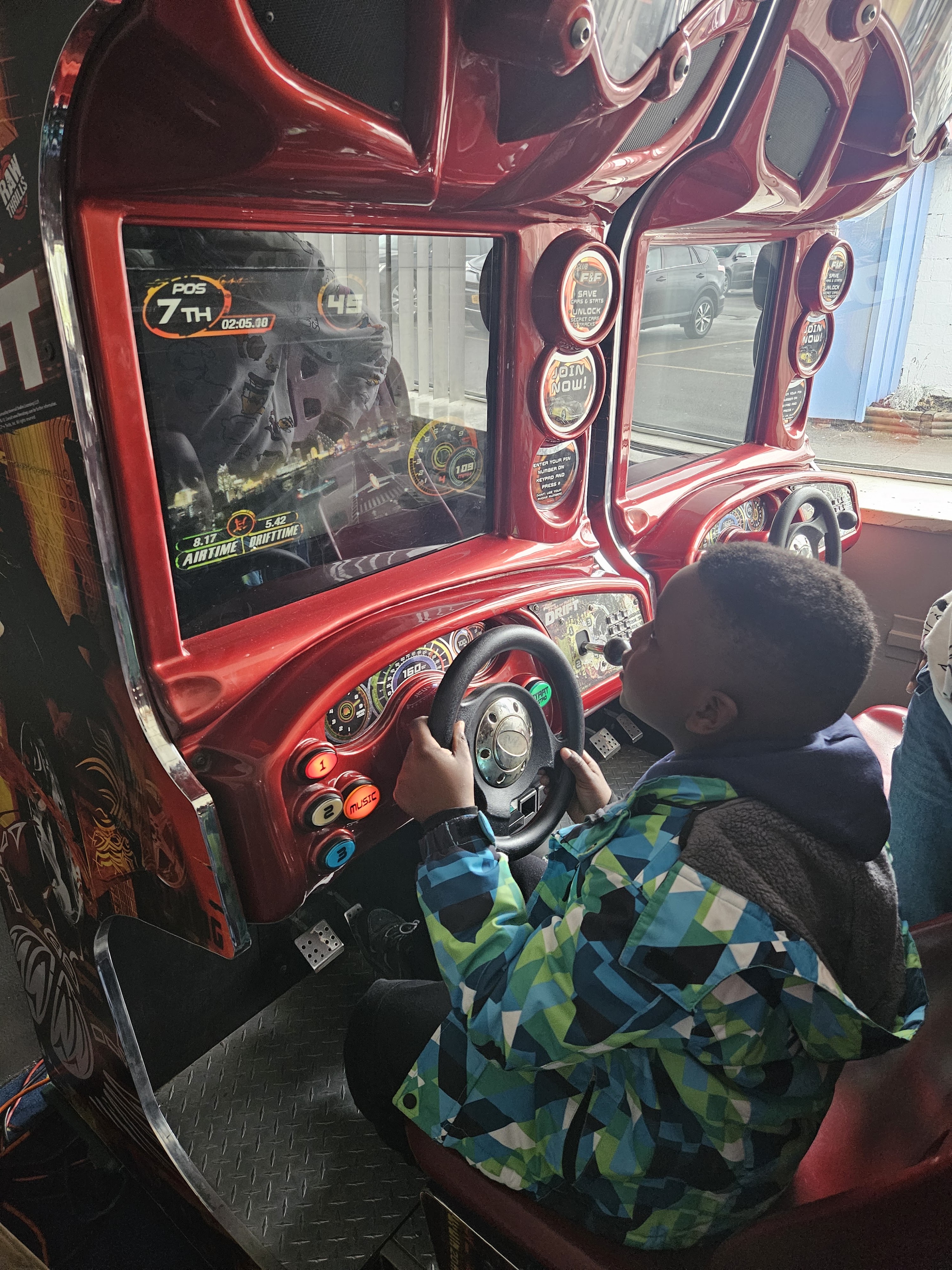 A child sits in a red racing arcade game, holding the steering wheel and looking at the screen.