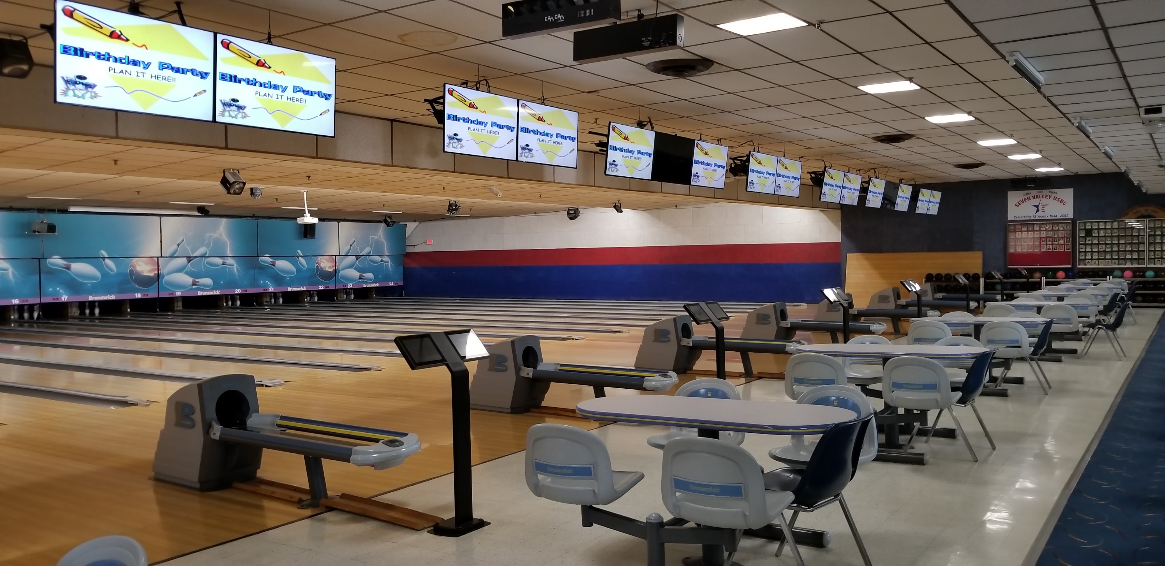 An empty bowling alley with multiple lanes, overhead scoring screens displaying birthday party promotions, and seating areas.