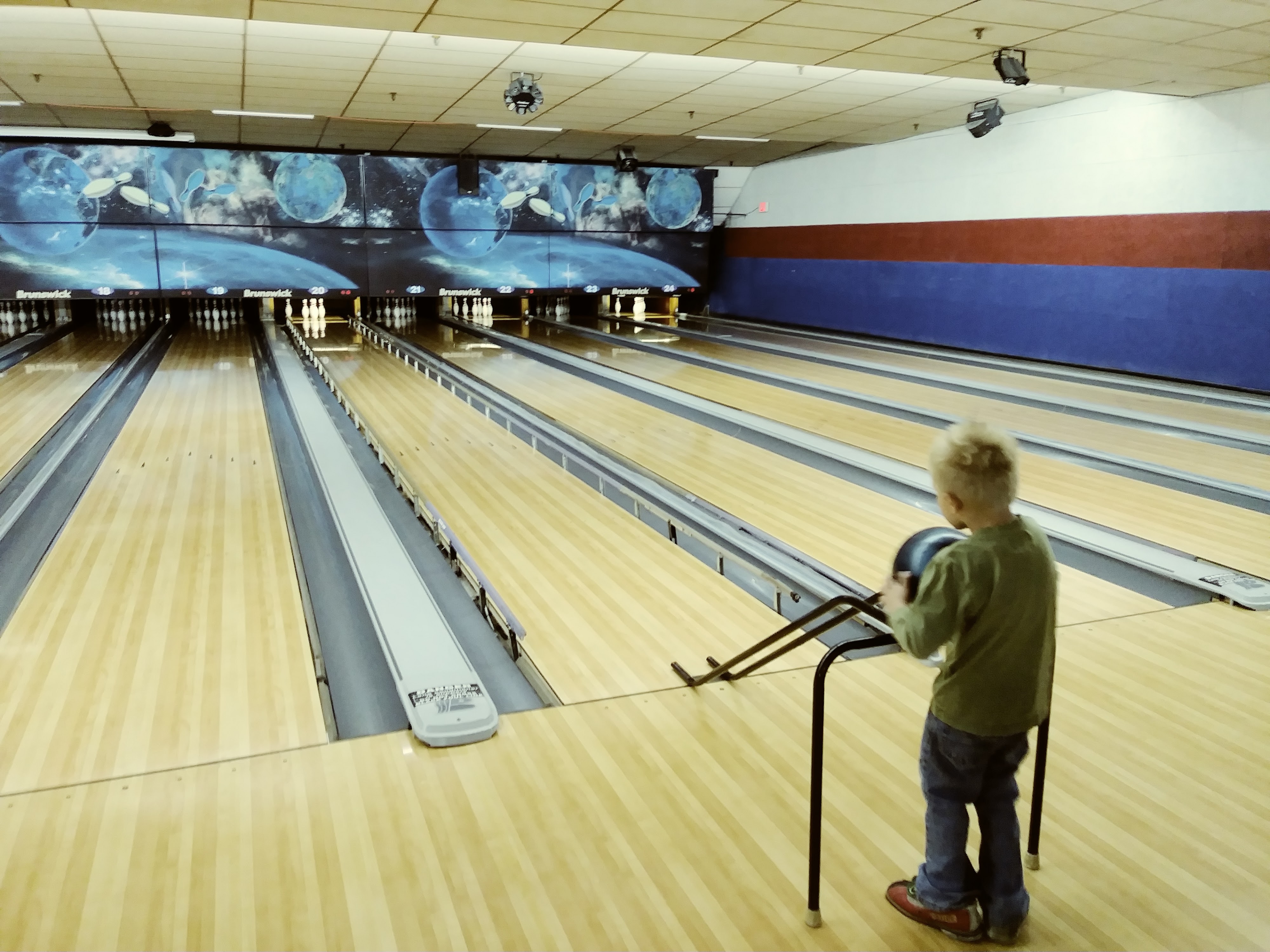 A young boy stands at the start of a bowling lane, holding a black bowling ball on a metal ramp, with multiple lanes stretching into the distance.