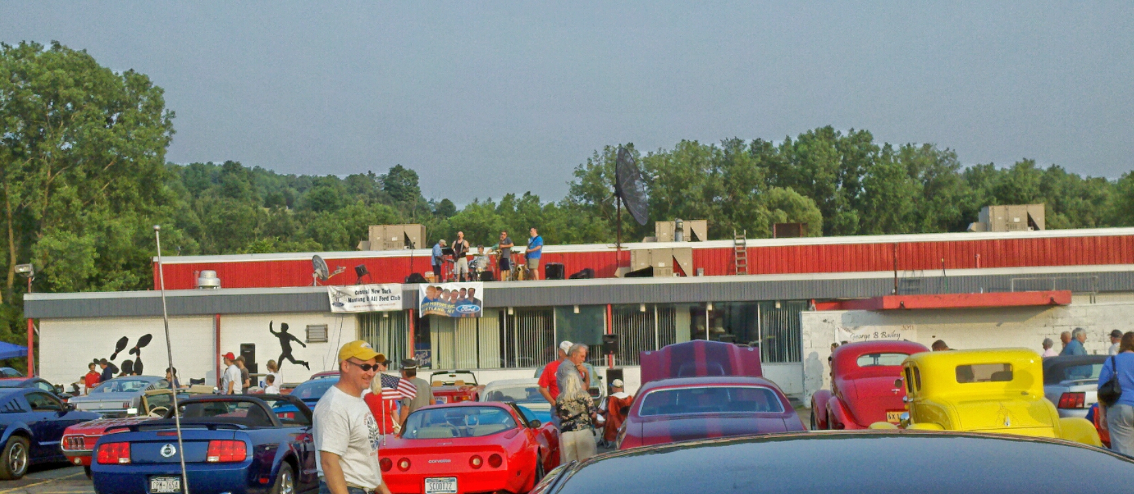 An outdoor car show features classic cars parked in front of a red-roofed building, with a band performing on the roof.