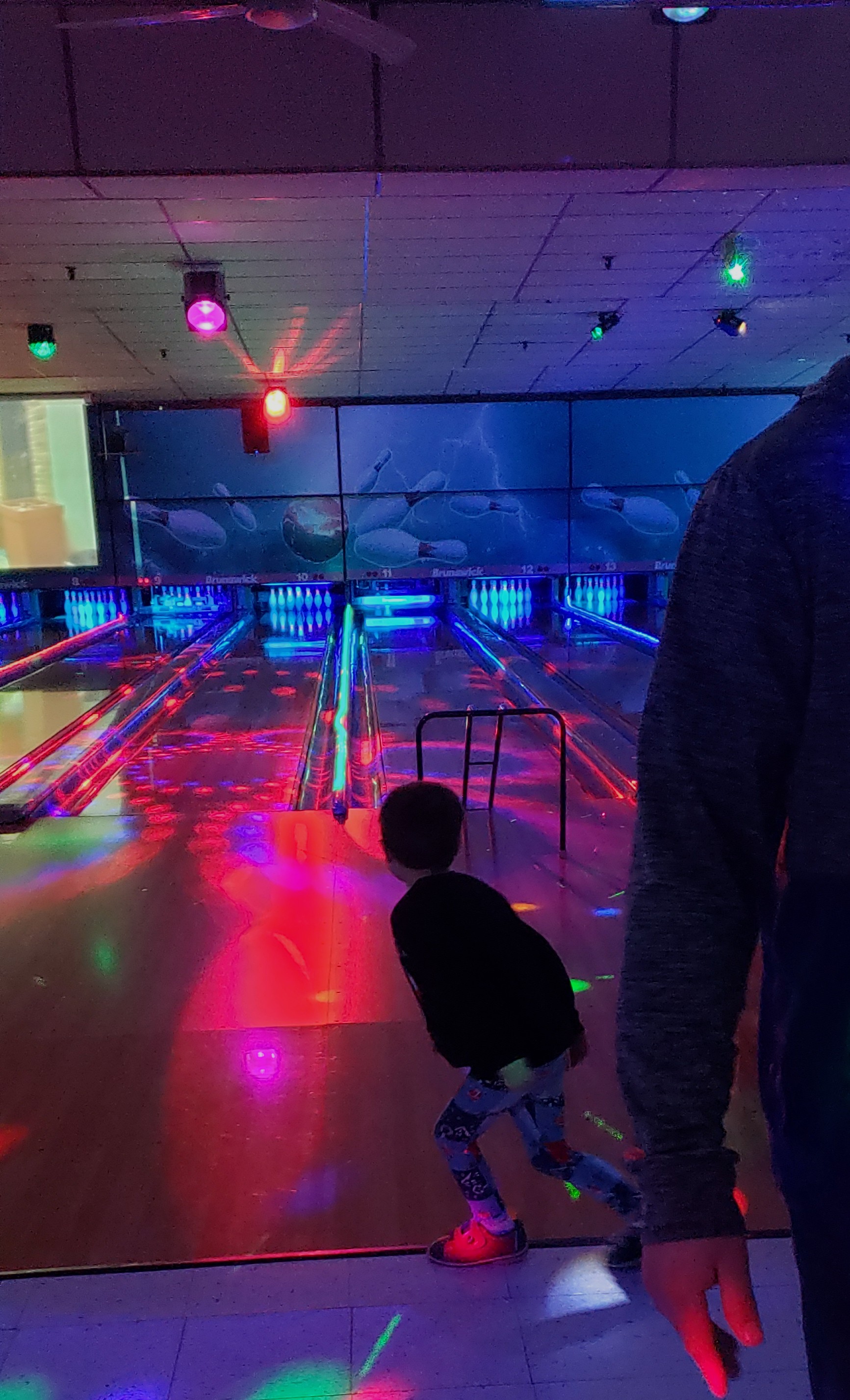 A child bowls in a dimly lit bowling alley, illuminated by red, blue, and green lights, with a bumper lane visible.
