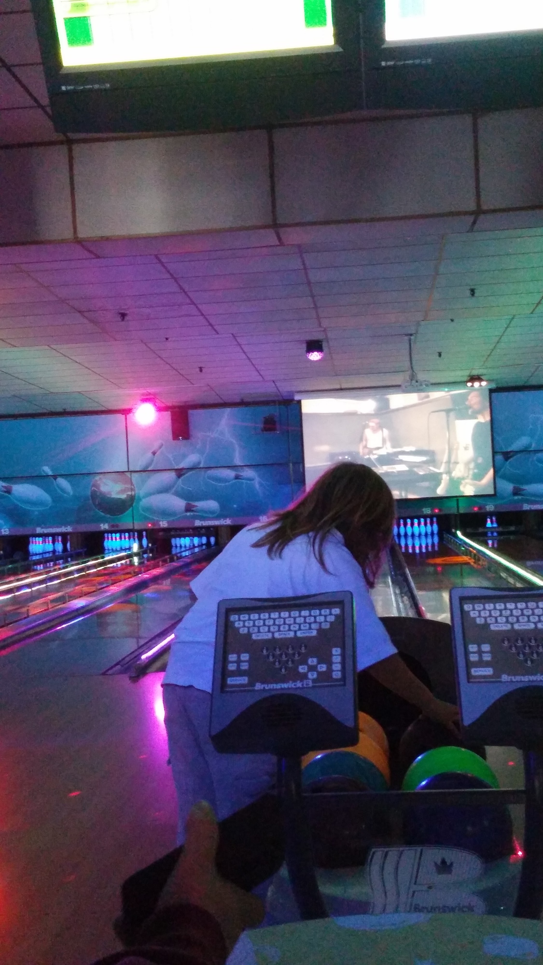 A person leans over a bowling ball return on a cosmic bowling lane with glowing pins and a large video screen in the background.