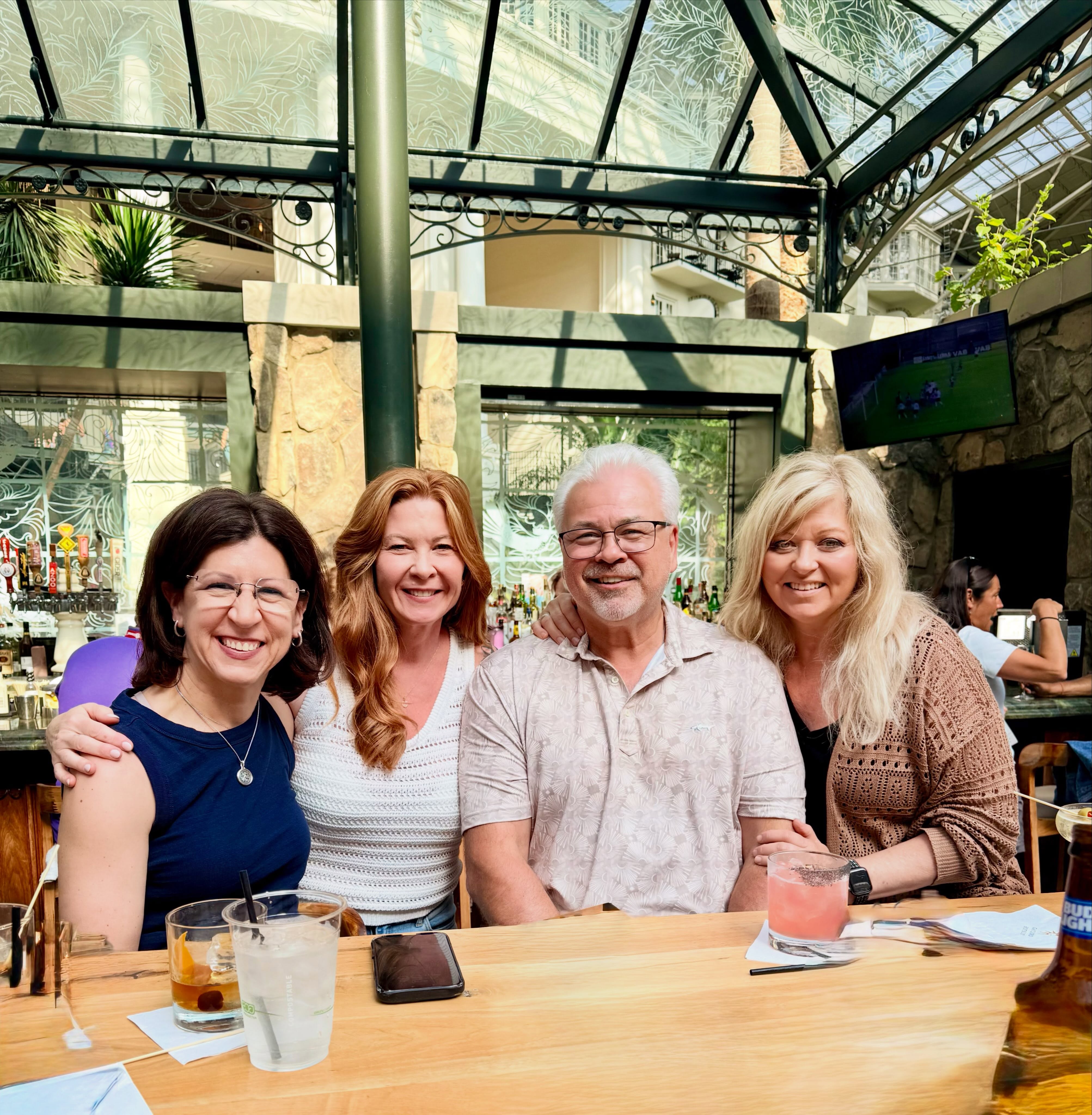 Four smiling adults sitting together at a bright, sun-drenched bar with a glass ceiling.