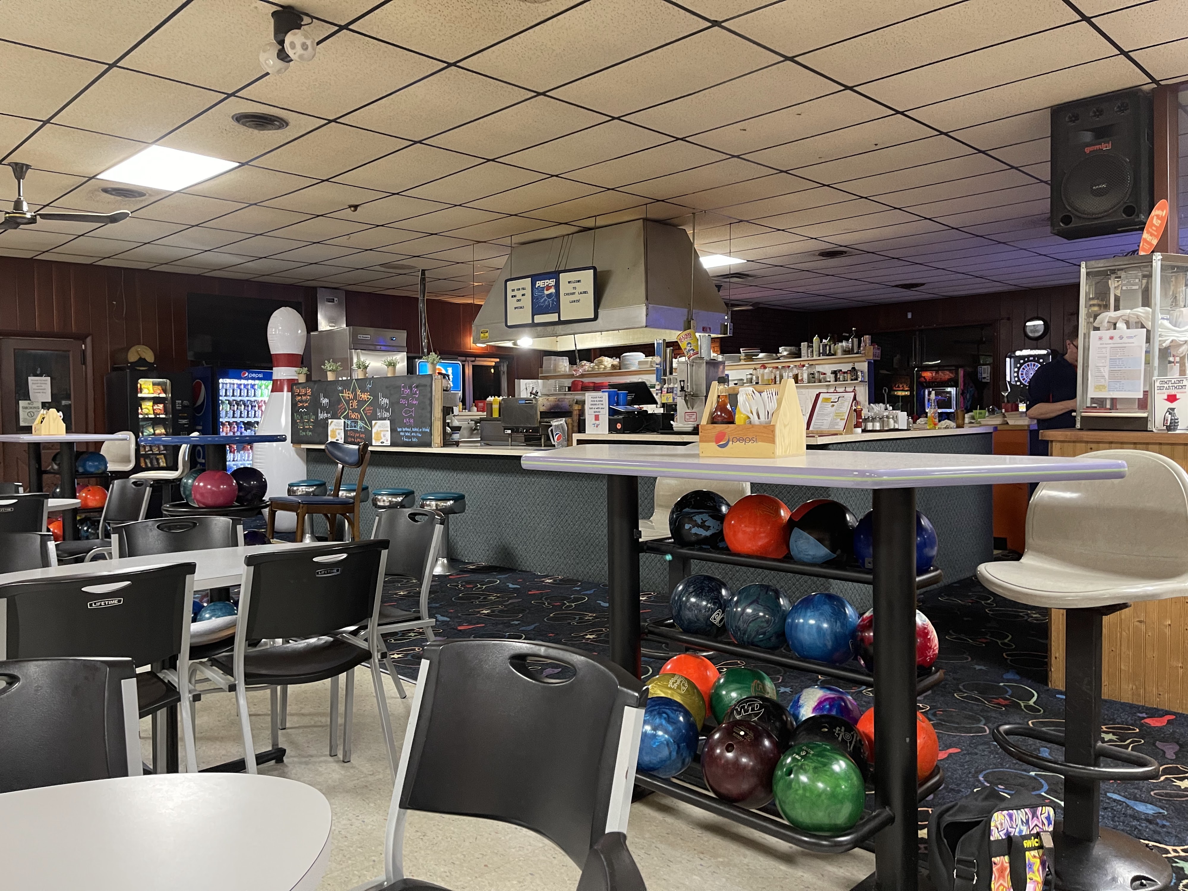The snack bar and house ball racks inside a bowling center with tables and chairs in the foreground.