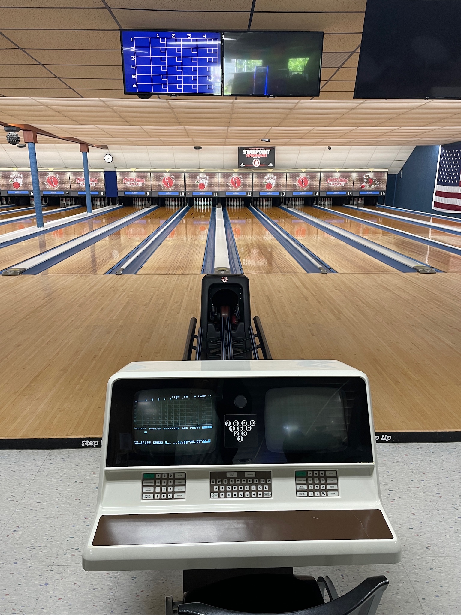 A view from behind a vintage scoring console looking down several polished bowling lanes at a bowling center.