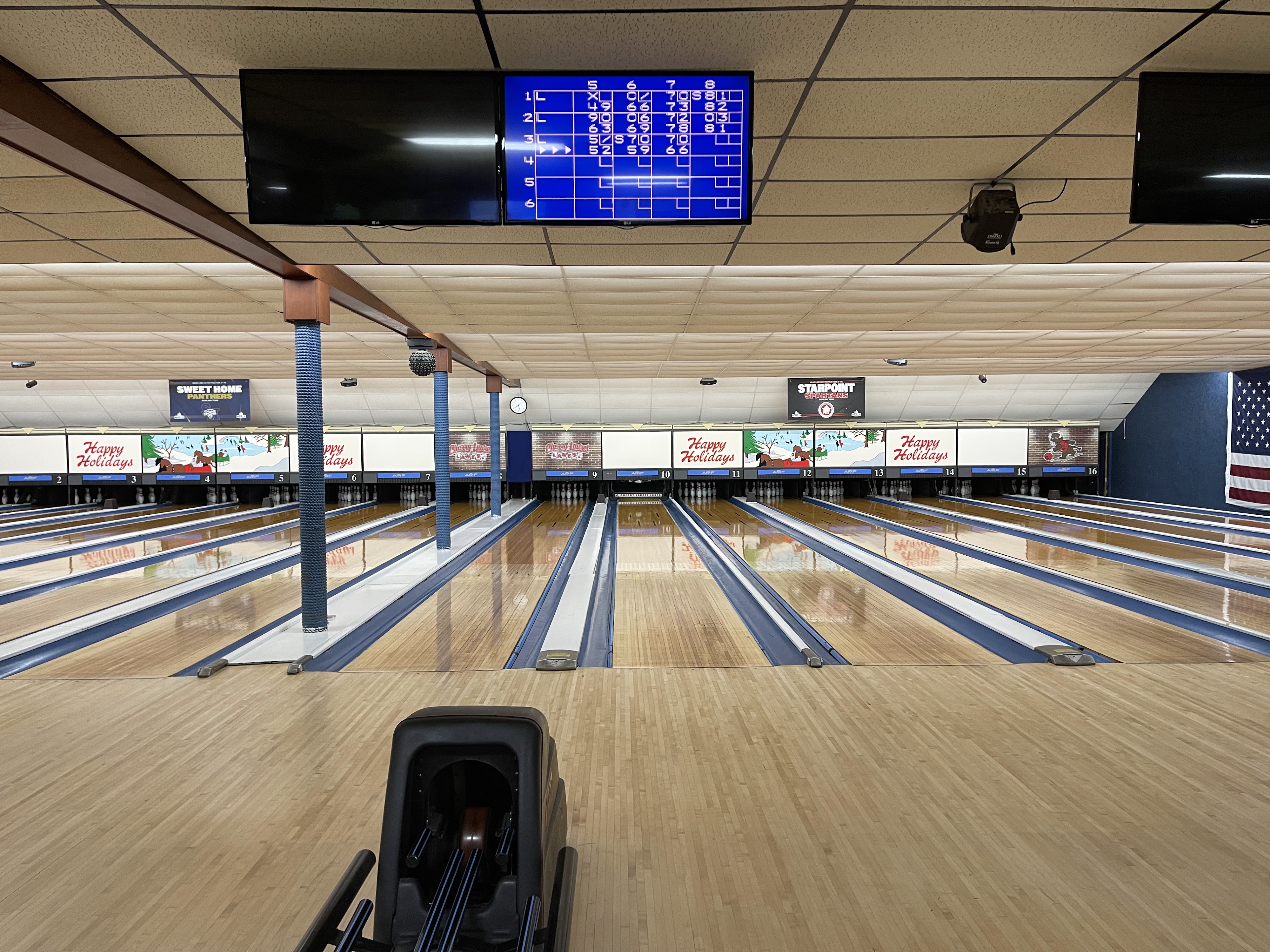A wide view of sixteen polished bowling lanes with active overhead scoring monitors and festive holiday decorations.