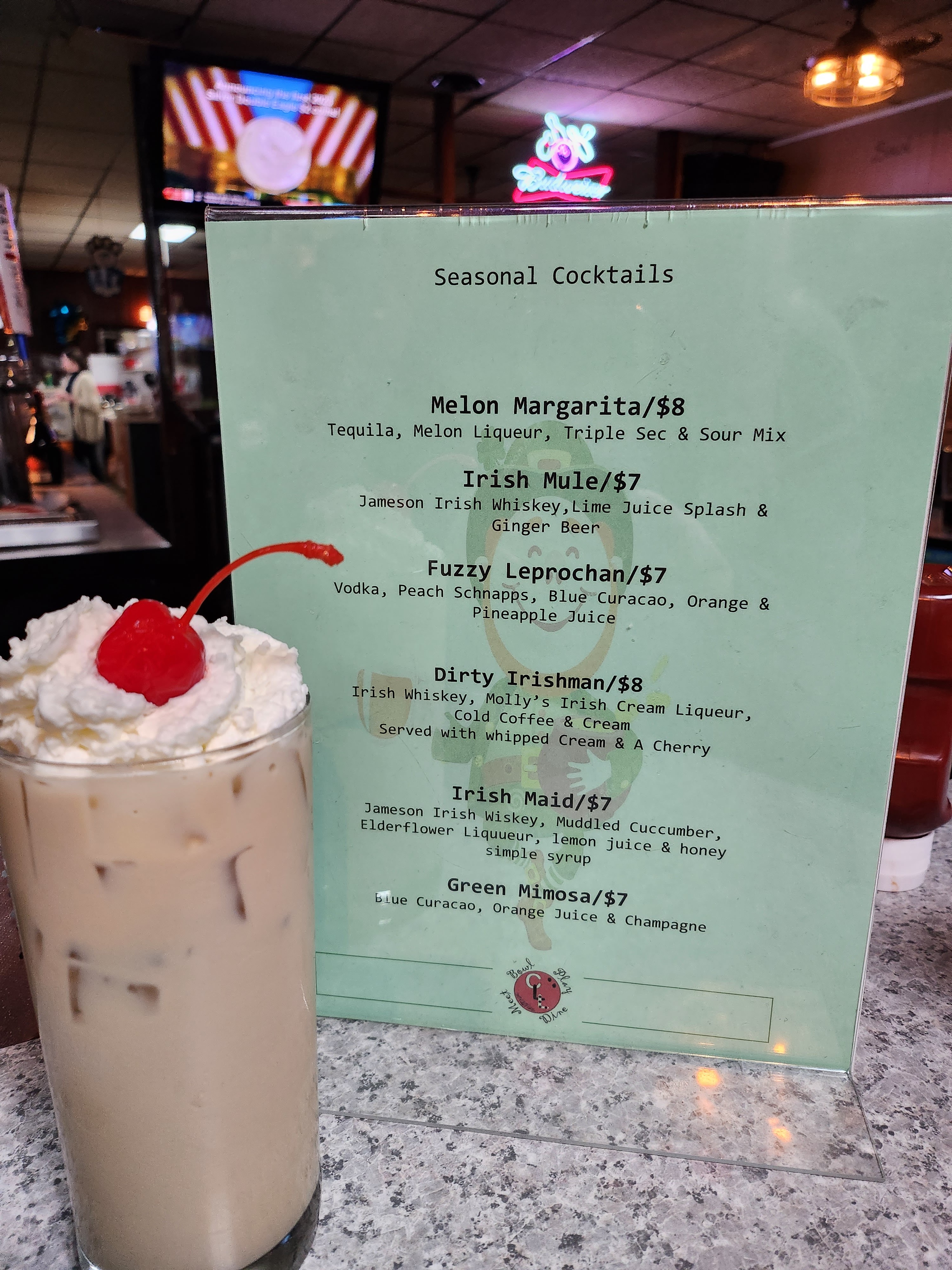 A tall iced cocktail with whipped cream and a cherry next to a green seasonal drink menu on a bar counter.