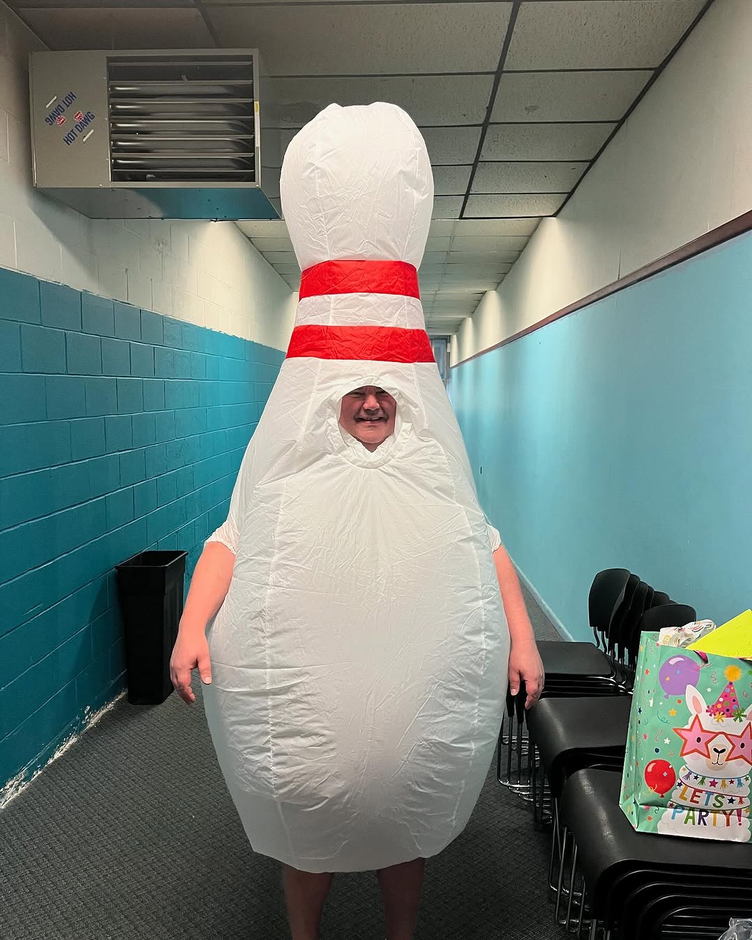 A man in a large inflatable bowling pin costume smiles in a blue hallway near a birthday gift bag.