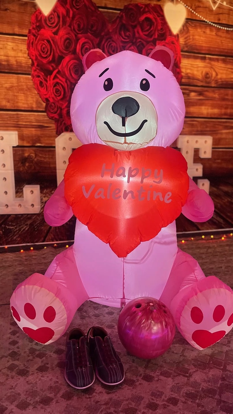 A large pink inflatable teddy bear holding a 'Happy Valentine' heart next to a bowling ball and shoes.