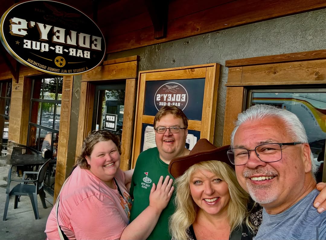 Four people smiling in a selfie outside of a building featuring a Bar-B-Que restaurant sign.