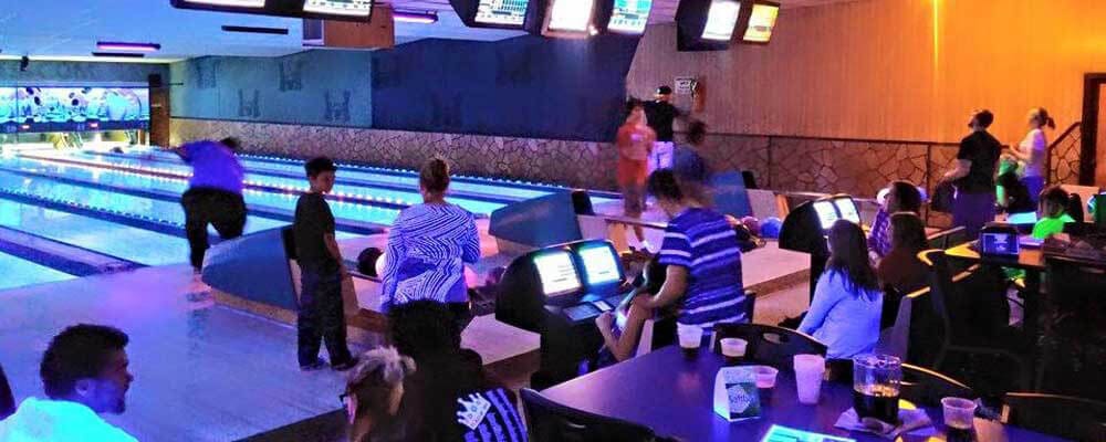 A wide shot of people bowling in a center with blue cosmic lighting and drinks on tables in the foreground.