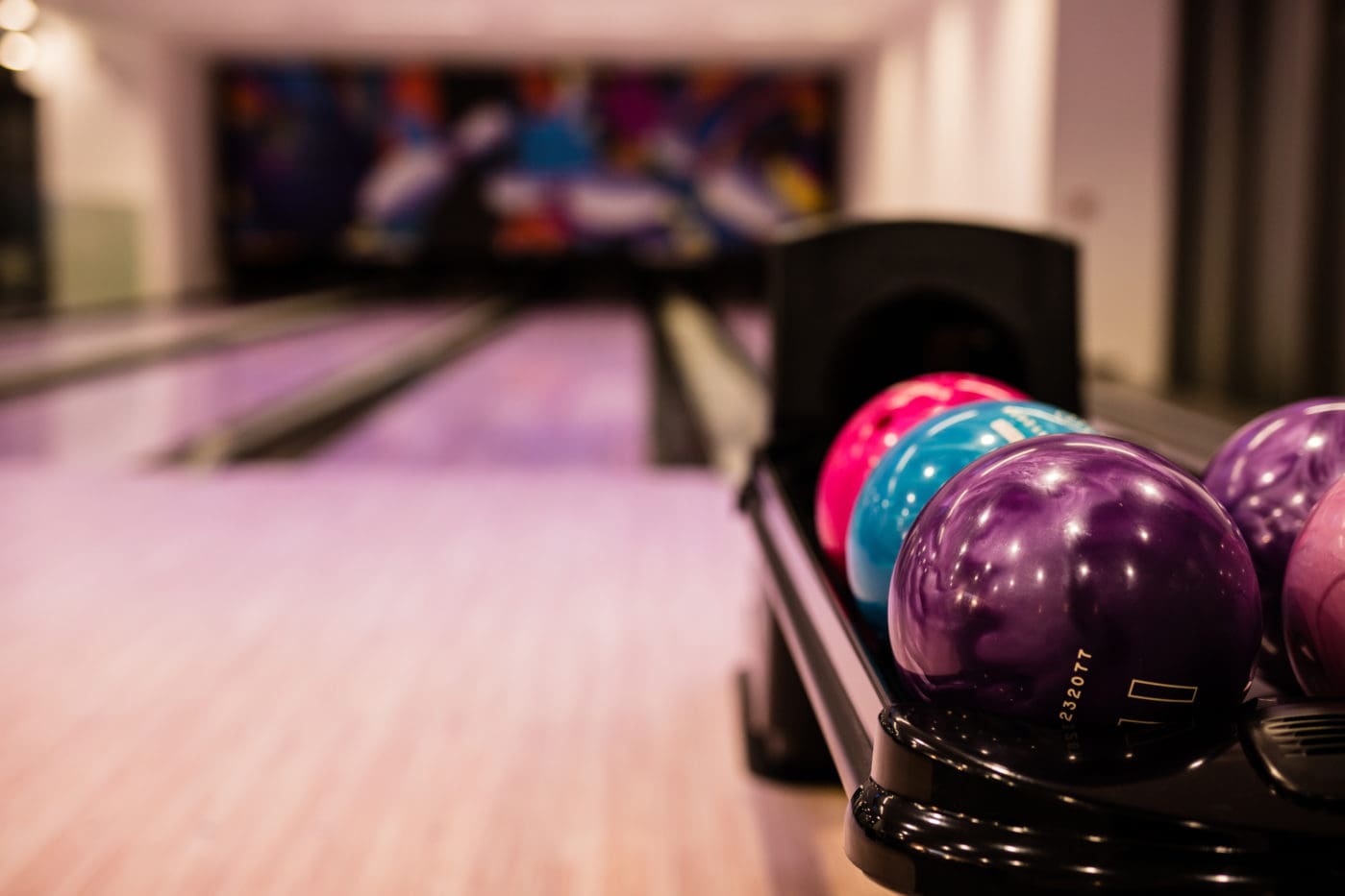 Close-up of colorful pink, blue, and purple bowling balls on a return rack with blurred lanes in the background.