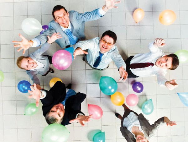 Six business professionals looking up and reaching out among colorful balloons on a tiled floor.