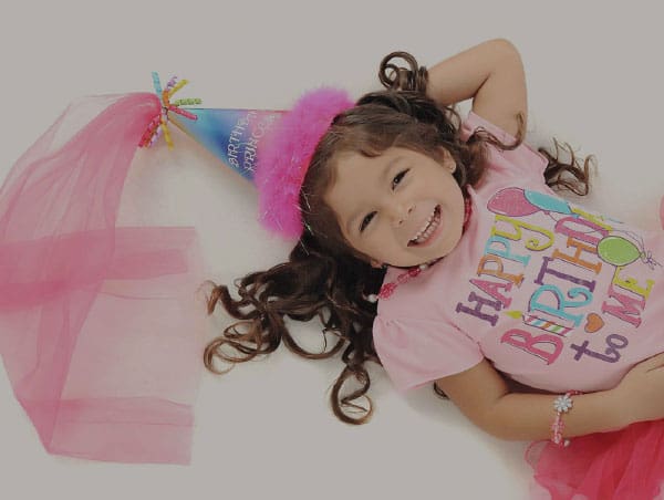 A young girl wearing a pink birthday shirt and a party hat smiles while lying down.