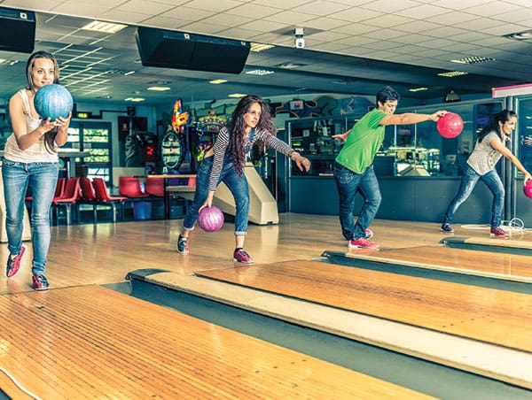 Four people bowling together on adjacent lanes in a brightly lit bowling center.