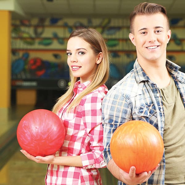 A man and a woman smiling while holding a red and an orange bowling ball at a bowling alley.