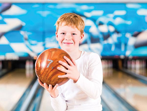 A smiling young boy with red hair holding an orange bowling ball in a bowling alley.