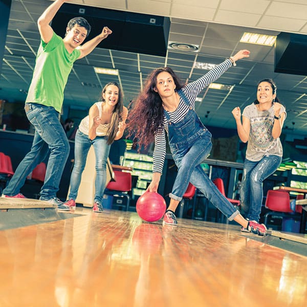 A woman in overalls rolls a pink bowling ball while three friends cheer behind her on a wooden lane.