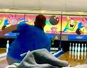 A bowler in a blue shirt mid-delivery on a lane with colorful cartoon graphics above the pins.