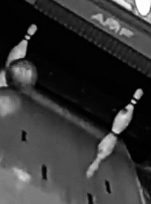 A black and white close-up image of a bowling ball hitting pins on a lane.