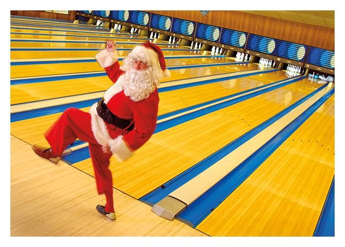 Santa Claus posing playfully on a bowling lane approach in front of several rows of pins.
