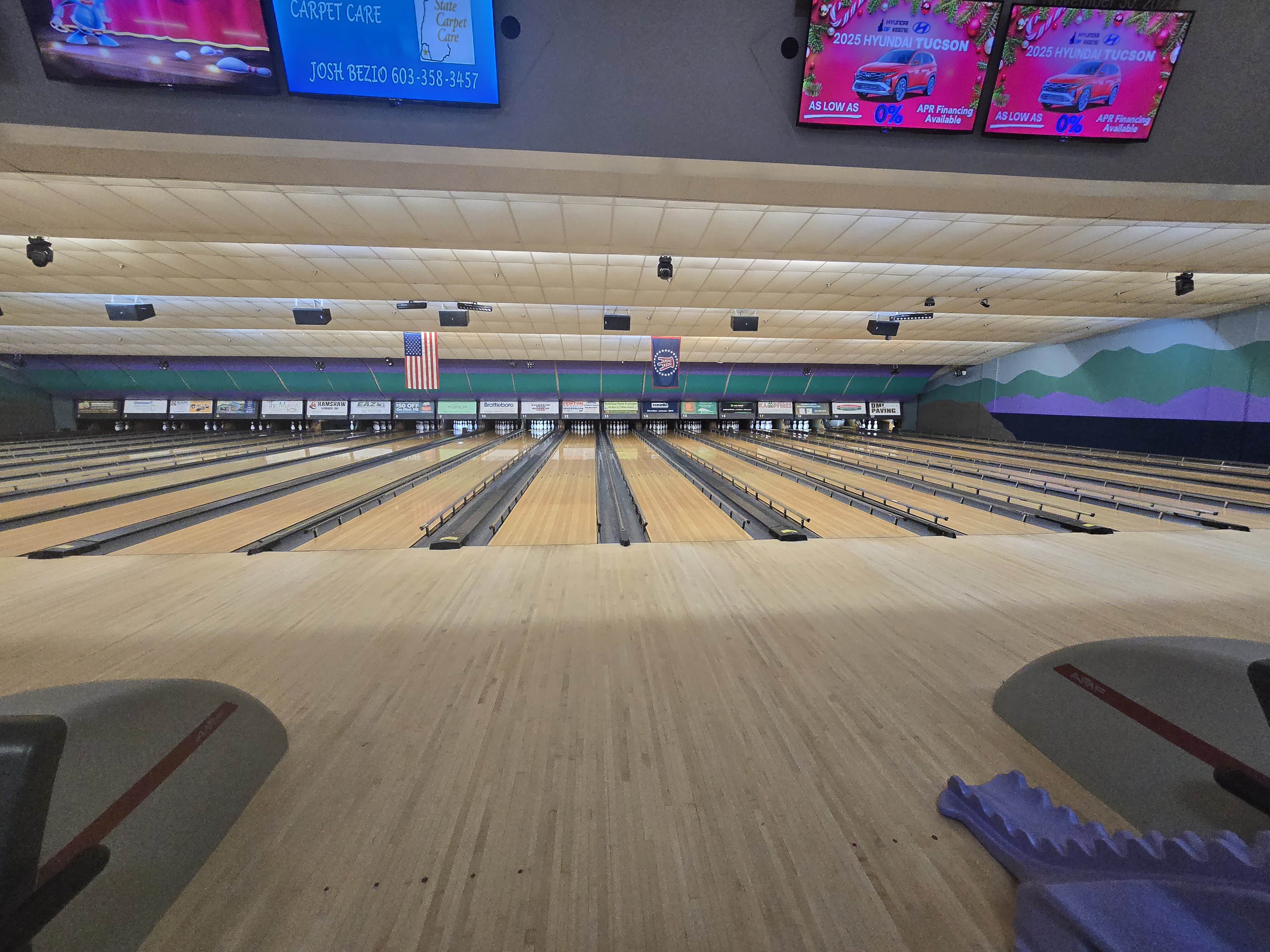 Wide view of numerous polished bowling lanes with overhead scoring monitors and a mountain mural on the back wall.